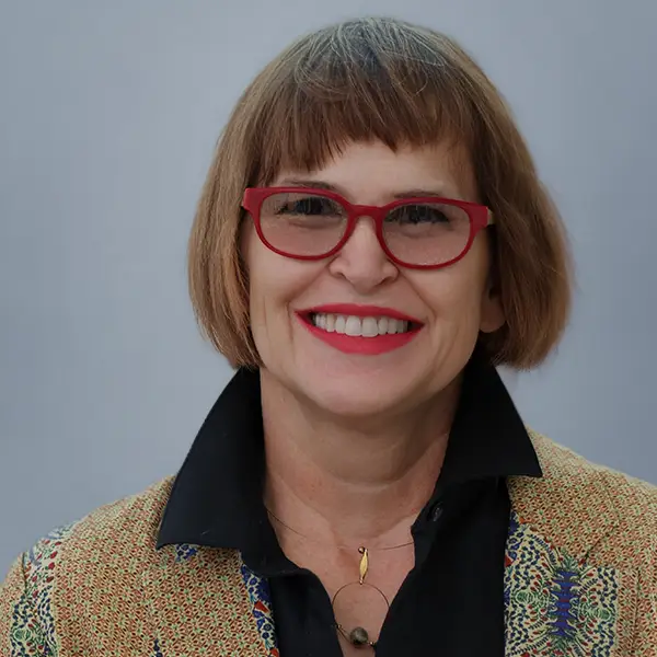 Smiling portrait of a woman with short brown hair and red glasses, wearing a patterned blazer and black shirt against a gray background.