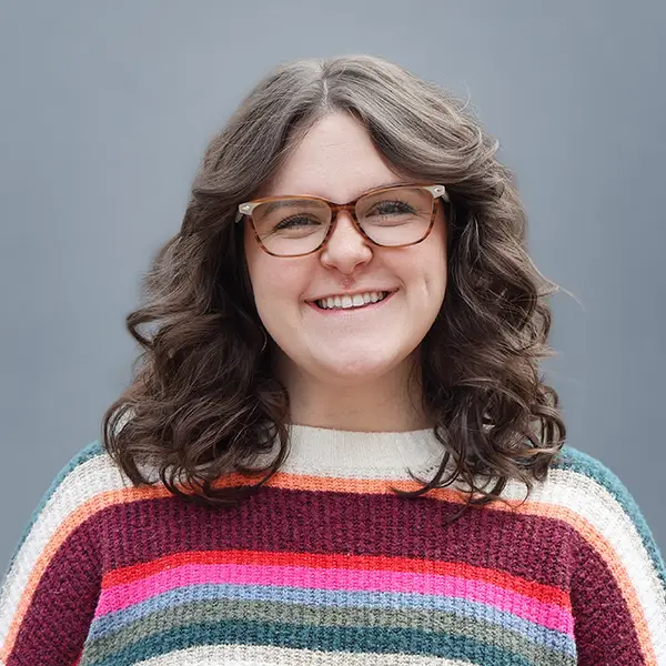 Smiling woman with wavy brown hair and glasses wearing a multicolored striped knit sweater against a gray backdrop.