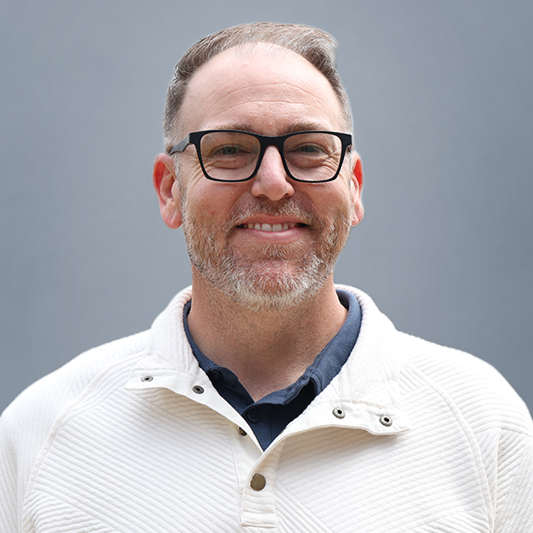 Portrait of a smiling man with dark-framed glasses, short gray hair and a light beard, wearing a white knit pullover against a gray backdrop.
