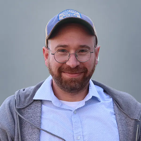 Smiling man with a beard and round glasses wearing a blue Tennessee Wesleyan cap and gray jacket outdoors.