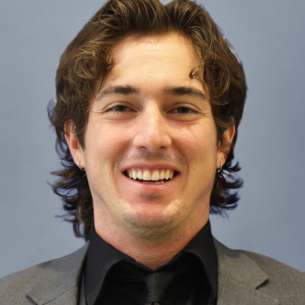 Smiling man in a gray suit and black shirt, posed against a blue background.