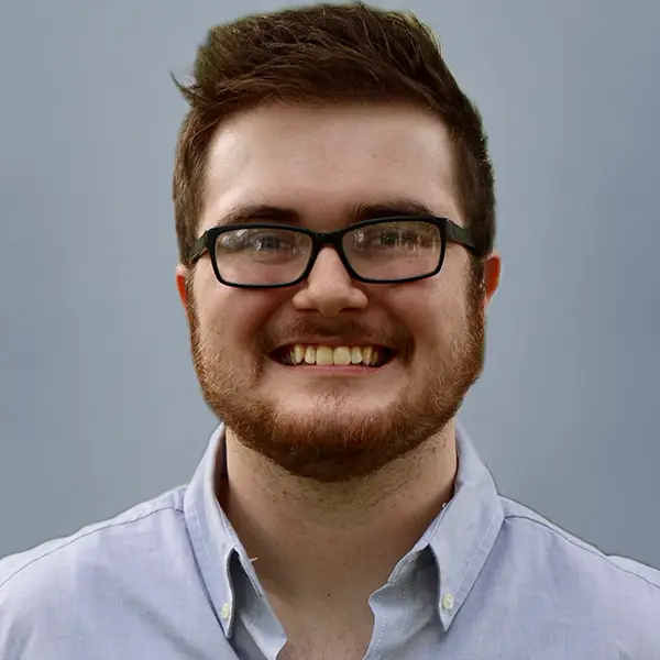 Smiling man with short brown hair and glasses, wearing a light blue button-down shirt against a gray background.