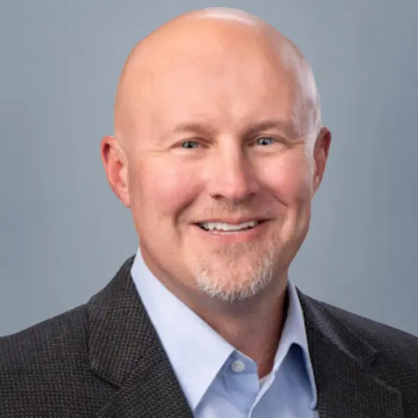 Professional headshot of a smiling bald man in a dark blazer and light blue shirt against a gray background.