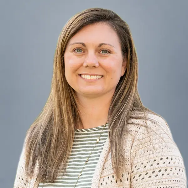 Portrait of a smiling woman with long light brown hair against a gray background, wearing a striped shirt and beige knitted cardigan.
