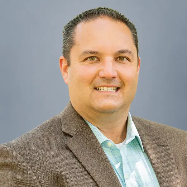 Smiling man in a brown blazer and light green plaid shirt posing for a headshot against a gray background