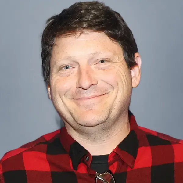 Smiling man with short brown hair wearing a red and black plaid shirt against a gray background.