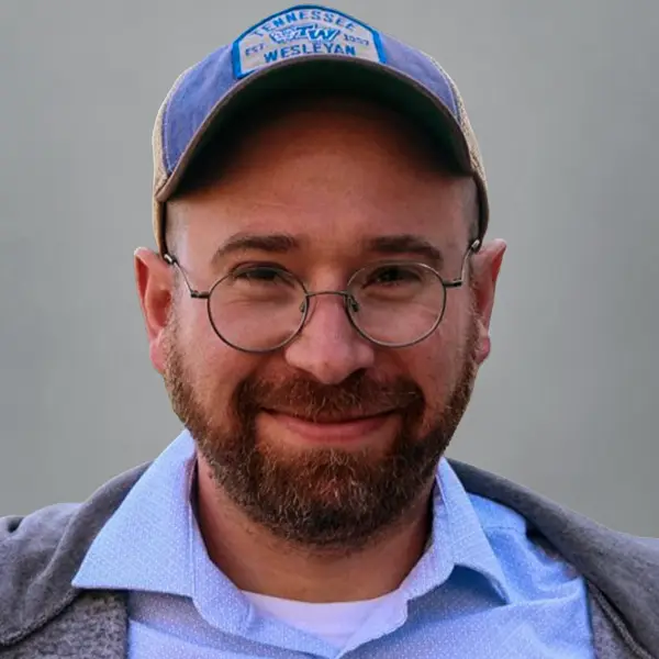Close-up portrait of a man with round glasses and a blue cap, smiling at the camera, with a beard and light blue shirt.