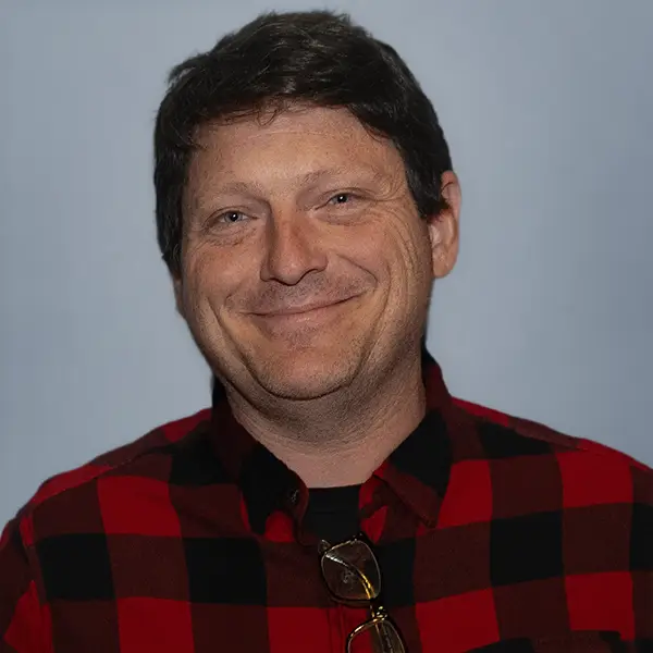 Portrait of a smiling man wearing a red-and-black plaid shirt against a light blue background.
