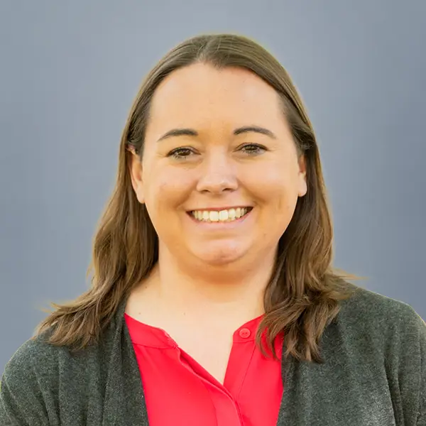 Smiling woman with shoulder‑length brown hair wearing a red blouse and gray cardigan, against a blue backdrop.