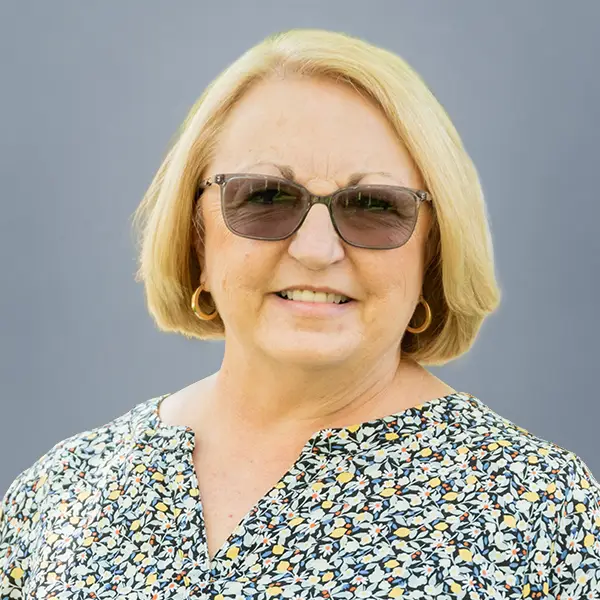 Smiling older woman with short blonde hair and sunglasses, wearing a patterned blouse and gold hoop earrings.