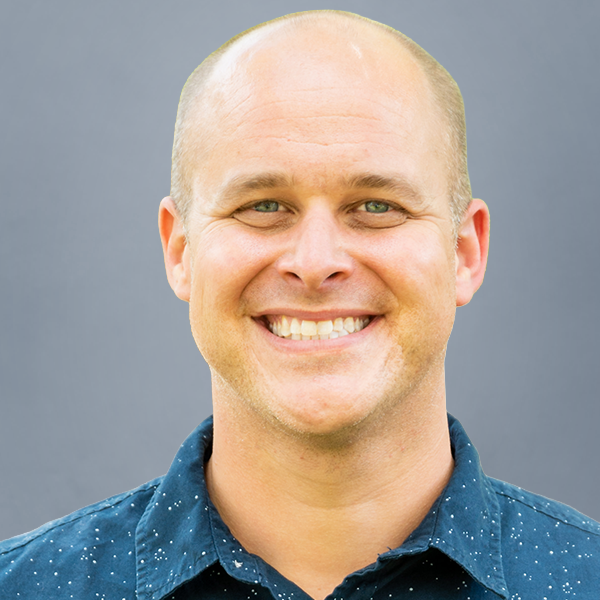 Smiling bald man facing the camera, wearing a blue button-up shirt, with a neutral gray background.