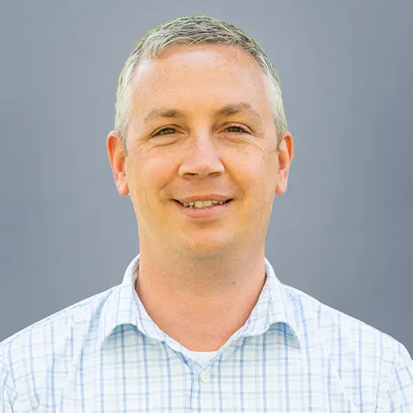 Portrait of a smiling man with short gray hair wearing a light blue plaid shirt against a gray background, facing the camera.