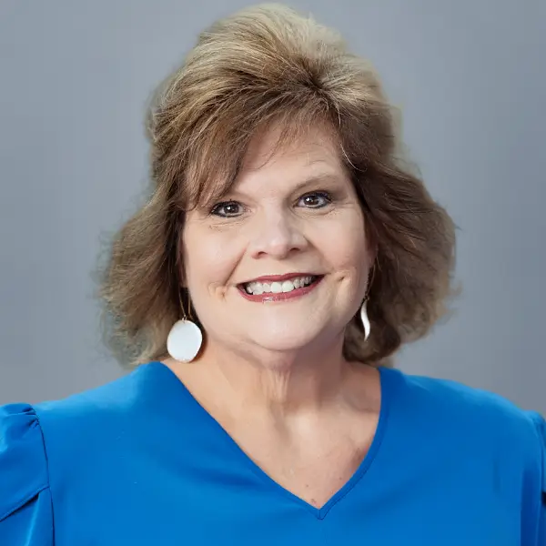 Smiling woman with short brown hair wearing a blue blouse and white drop earrings, facing the camera.