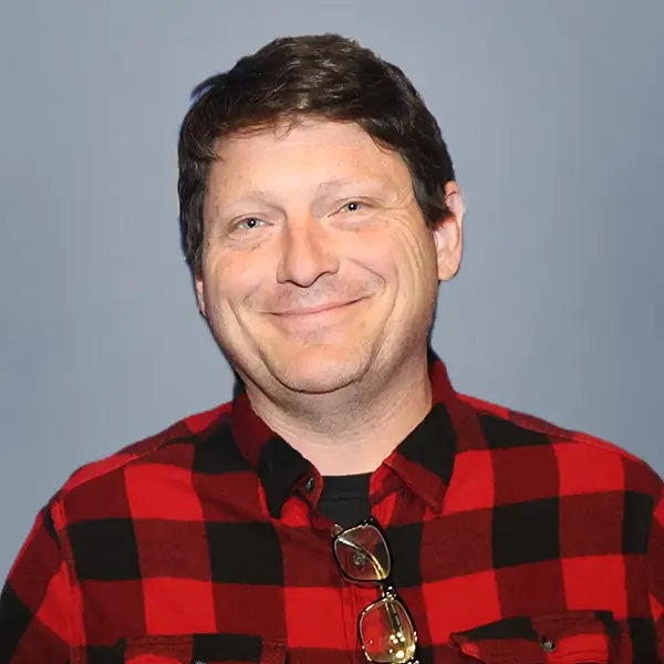 Smiling man with short dark hair wearing a red and black plaid shirt; glasses hang from his shirt and a blue-gray background is visible behind him.