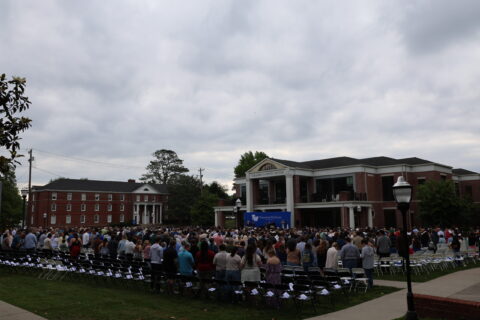 Large crowd at an outdoor university graduation ceremony, rows of chairs facing a stage on campus with a cloudy sky ahead of brick buildings.