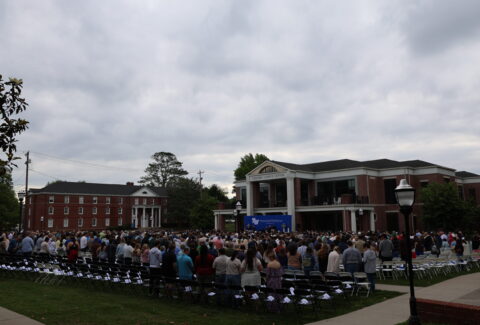 Large crowd at an outdoor university graduation ceremony, rows of chairs facing a stage on campus with a cloudy sky ahead of brick buildings.