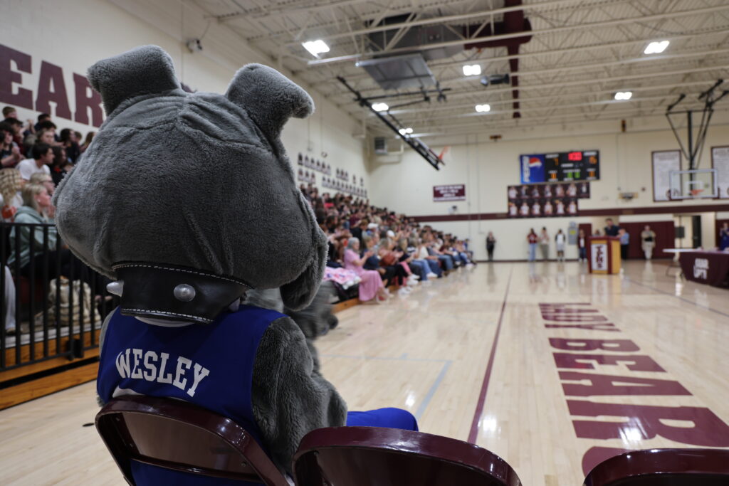 Mascot seated courtside facing a full gym, wearing a blue vest that reads 'WESLEY' with spectators and a scoreboard in the background