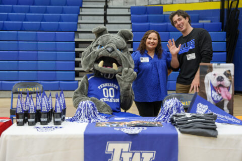 Tennessee Wesleyan Bulldogs mascot poses with two visitors at a gym table, blue banners and bleachers in the background.