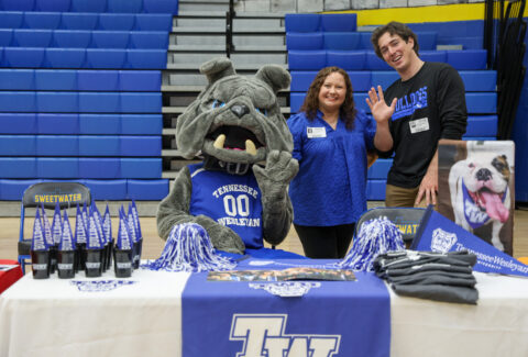 Tennessee Wesleyan Bulldogs mascot poses with two visitors at a gym table, blue banners and bleachers in the background.