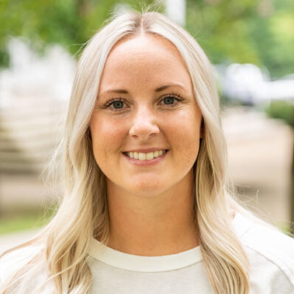 Smiling woman with long blonde hair outdoors, with greenery in the blurred background.