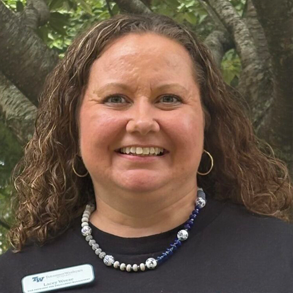 Portrait of a smiling woman with curly brown hair, wearing a beaded necklace and hoop earrings, outdoors.