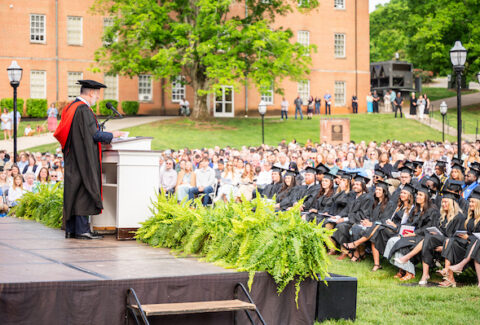 Speaker at a lectern delivering a commencement speech to graduates in caps and gowns outdoors on a university lawn with a brick building in the background.