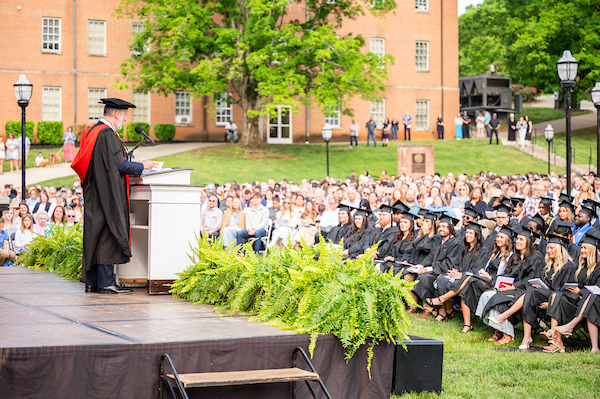 Speaker at a lectern delivering a commencement speech to graduates in caps and gowns outdoors on a university lawn with a brick building in the background.