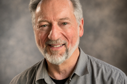 Smiling older man with gray hair and beard in a gray button-down shirt, posed against a neutral studio backdrop.