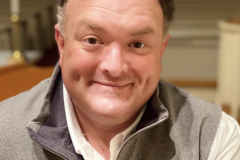 Close-up of a smiling man wearing a gray North Face fleece and a white shirt outdoors? indoors, cropped at shoulders.