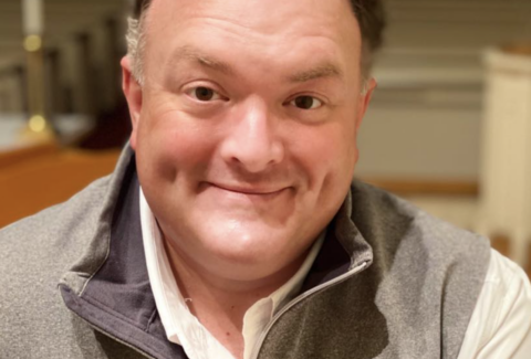 Close-up of a smiling man wearing a gray North Face fleece and a white shirt outdoors? indoors, cropped at shoulders.