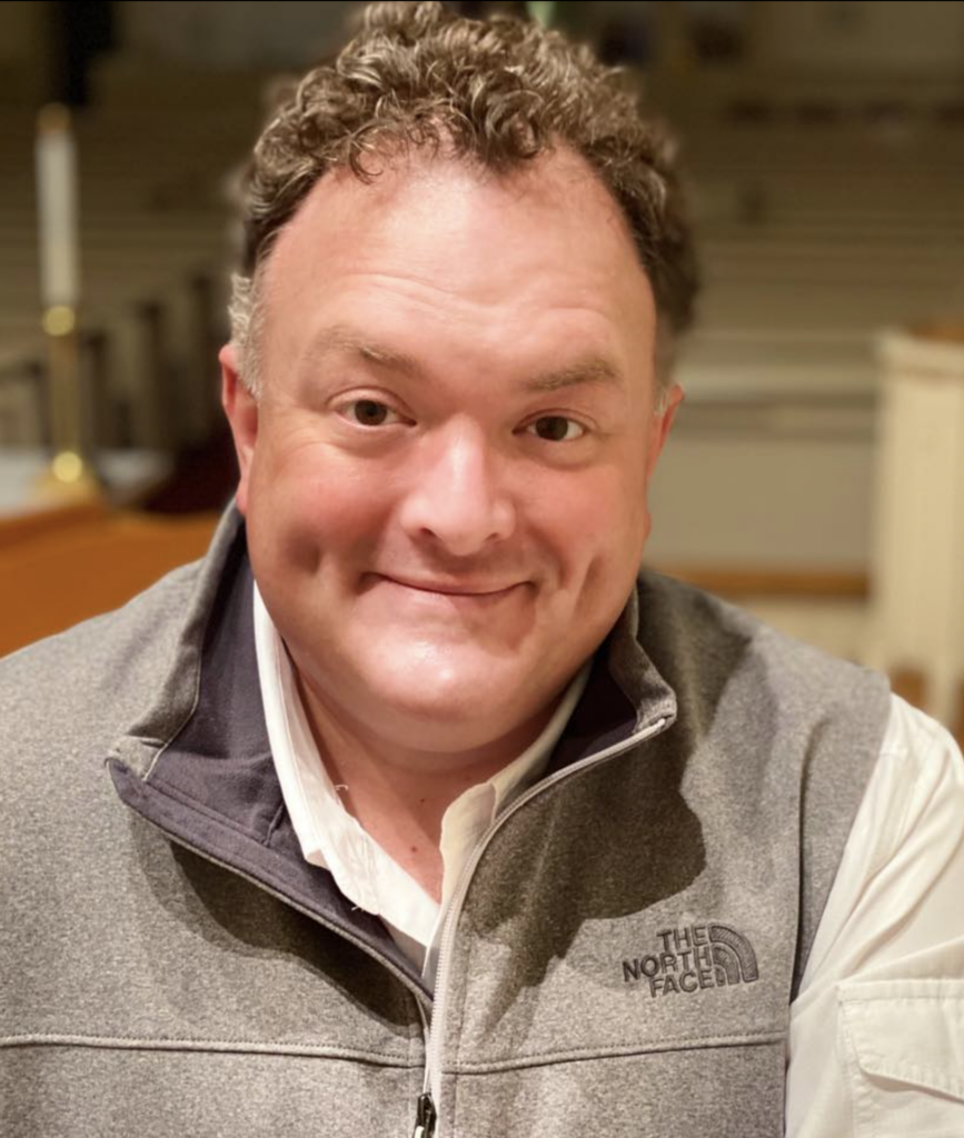 Close-up of a smiling man wearing a gray North Face fleece and a white shirt outdoors? indoors, cropped at shoulders.