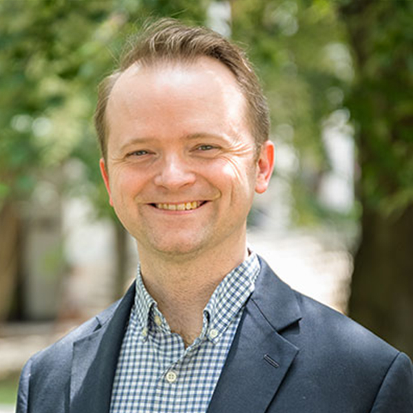 Smiling man in a navy blazer and checkered shirt photographed outdoors with greenery in the background.