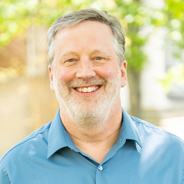 Smiling middle-aged man in a blue shirt outdoors, with a bright, natural background.