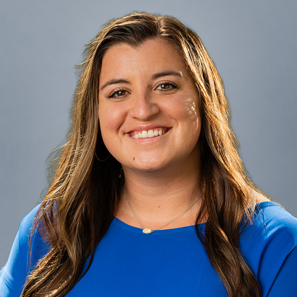 Smiling woman with long brown hair wearing a blue top against a gray backdrop.