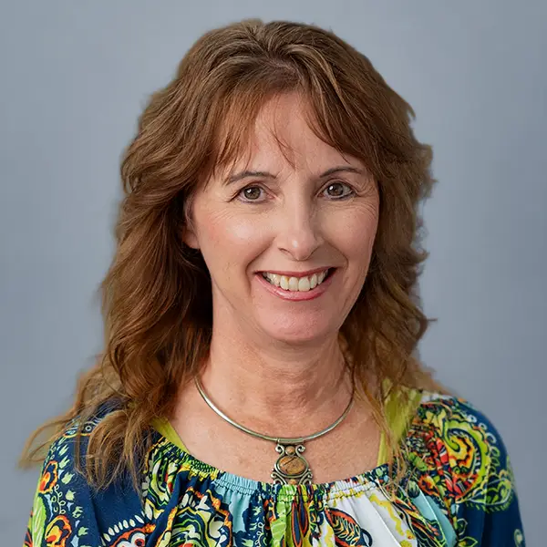 Smiling woman with wavy brown hair wearing a colorful patterned blouse and a silver necklace against a pale blue background.