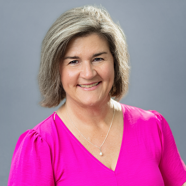 Smiling woman with short gray hair wearing a bright pink blouse and a pearl necklace against a gray backdrop.