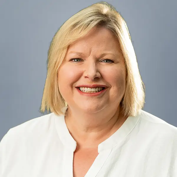 Smiling woman with short blond hair wearing a white blouse against a gray-blue background (head-and-shoulders portrait).