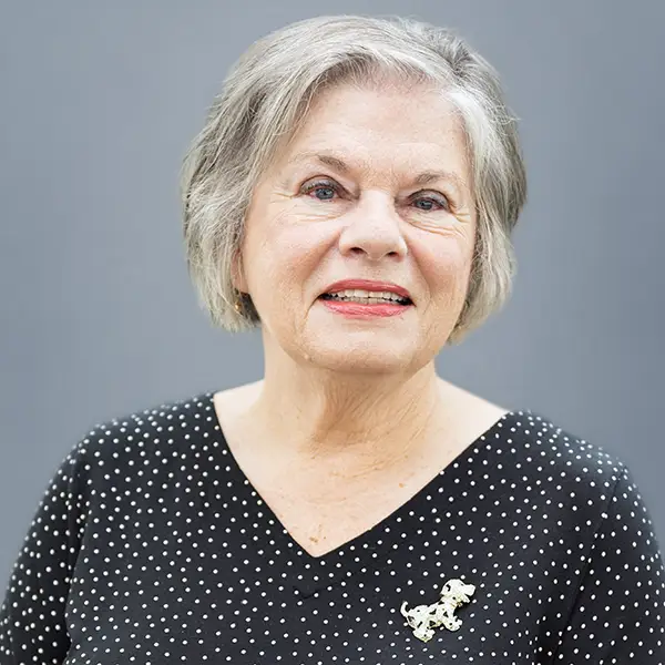 Portrait of an older woman with short gray hair and a gentle smile, wearing a black polka-dot blouse with a gold brooch against a gray background.