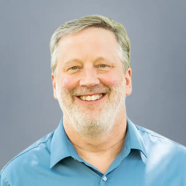 Portrait of a smiling middle-aged man with gray hair and beard, wearing a blue shirt against a gray backdrop.