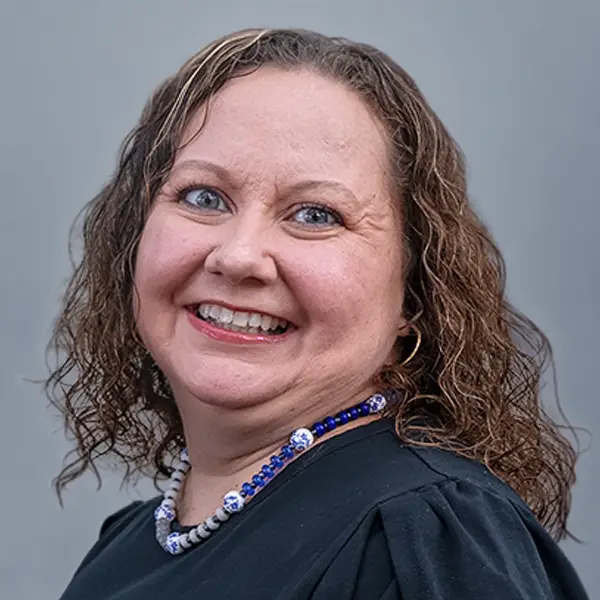 Portrait of a smiling woman with shoulder-length curly brown hair, wearing a blue beaded necklace and dark top against a gray backdrop.
