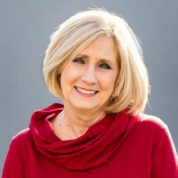 Smiling blonde woman in a red sweater against a neutral gray background.