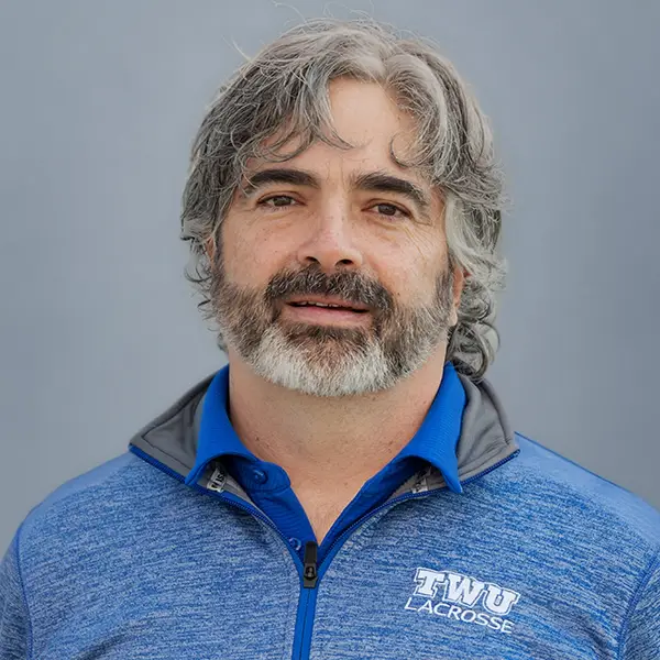 Portrait of a middle-aged man with gray hair and beard wearing a blue TWU Lacrosse jacket against a neutral background.