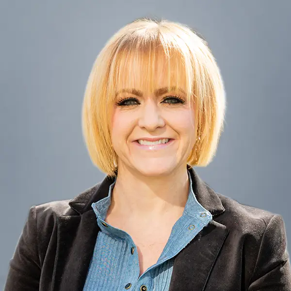 Smiling blonde woman in a black blazer and denim shirt against a neutral background.