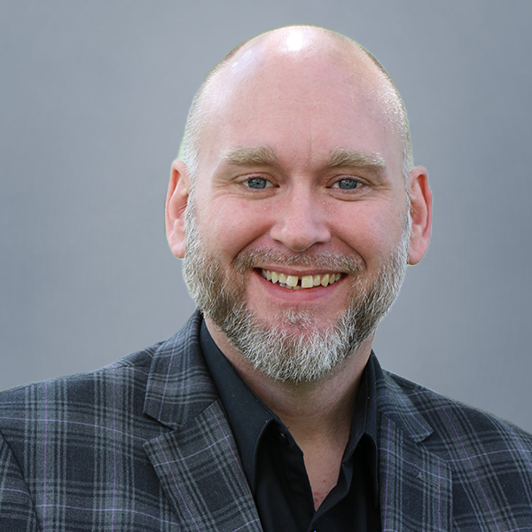 Smiling bald man with a gray beard wearing a dark plaid blazer, neutral gray background.