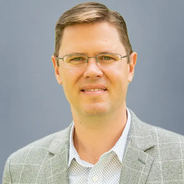 Professional headshot of a man wearing glasses and a light gray checked blazer, against a plain gray background.