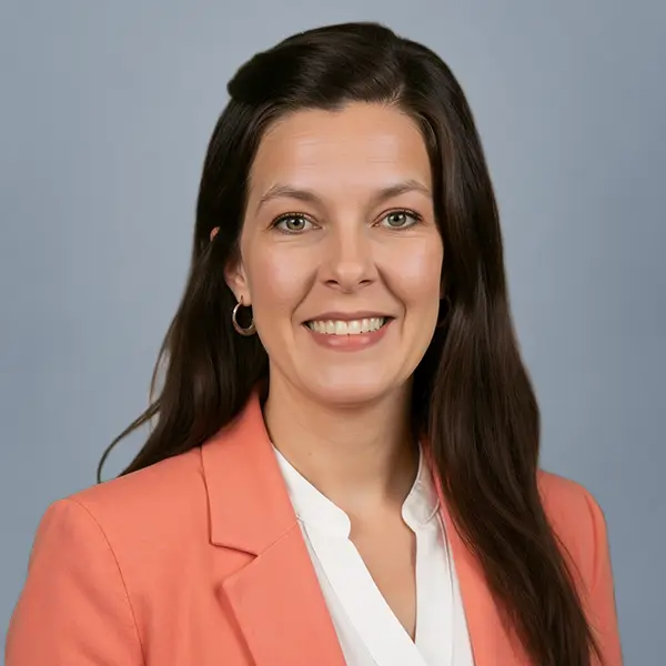 Smiling woman with long dark hair in a peach blazer and white blouse, against a gray backdrop.