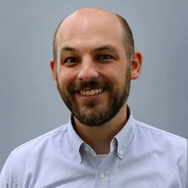 Smiling man with a beard and a light blue button-down shirt against a gray background.