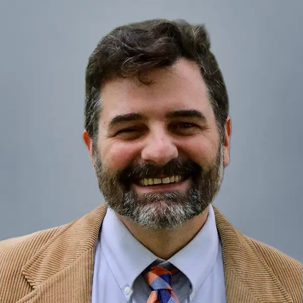 Smiling man with dark hair and beard in a tan blazer, light blue shirt, and patterned tie against a gray background (professional headshot).