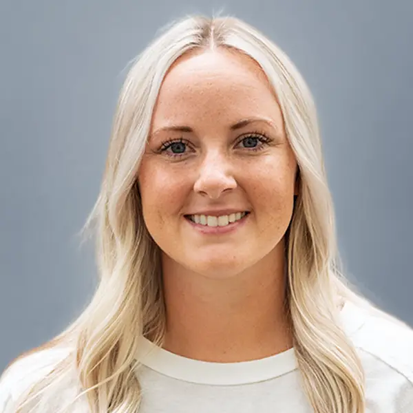 Head-and-shoulders portrait of a smiling woman with long blonde hair against a gray background, wearing a light top.