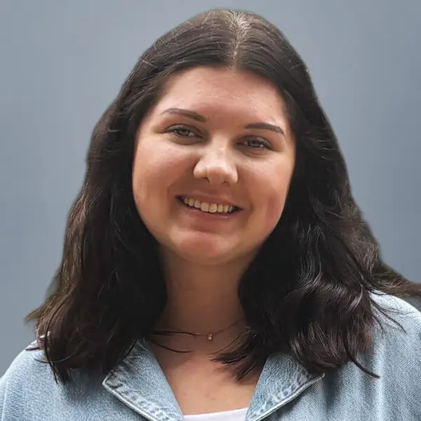 Portrait of a smiling woman with shoulder-length dark hair wearing a light denim jacket against a neutral blue-gray background.